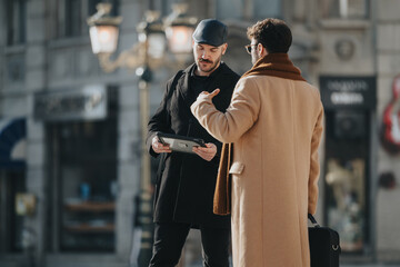 Two stylish men engaging in conversation on a city street with digital tablet.