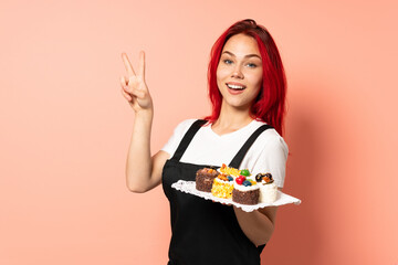 Pastry chef holding a muffins isolated on pink background smiling and showing victory sign