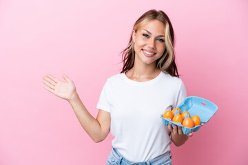 Young Russian woman holding eggs isolated on pink background extending hands to the side for inviting to come