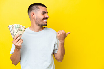 Young caucasian man taking a lot of money isolated on yellow background pointing to the side to present a product