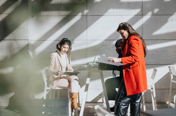 Focused female people collaborating in a bright workspace with shadows. One woman reviews documents while the other listens to music and takes notes.