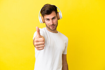 Young caucasian handsome man isolated on yellow background listening music and with thumb up