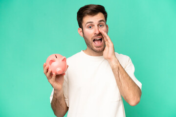 Young caucasian man holding piggybank isolated on green background shouting with mouth wide open