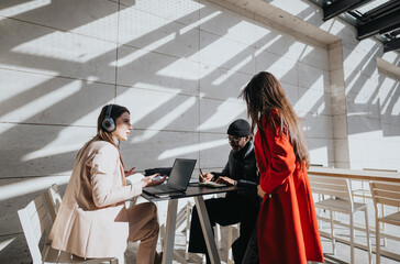 Young people engage in a productive business meeting, utilizing laptops and headphones at an outdoor cafe with sunlight casting geometric shadows.