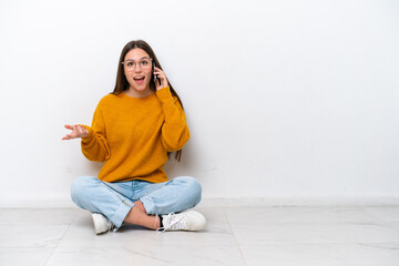 Young girl sitting on the floor isolated on white background keeping a conversation with the mobile phone with someone