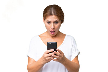 Fototapeta premium Middle-aged caucasian woman over isolated background looking at the camera while using the mobile with surprised expression