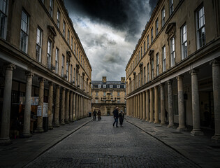 Fototapeta premium Dramatic urban street scene with historic architecture, tall columns, and moody overcast skies, as a few pedestrians walk along the cobblestone pathway.
