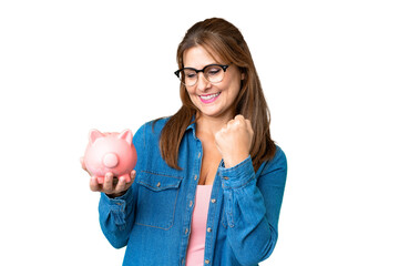 Middle age caucasian woman holding a piggybank over isolated background celebrating a victory