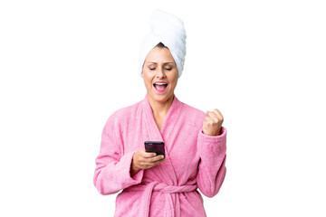 Middle age caucasian woman in a bathrobe over isolated background with phone in victory position