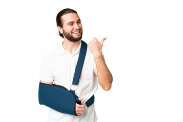 Young handsome man with broken arm and wearing a sling over isolated chroma key background pointing to the side to present a product
