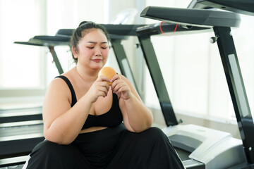 Chubby Asian woman exercise in gym, female contemplates her snack, reflecting on dietary choices in fitness journey. moment of thought, examines Hamburger, considering health and wellness goals