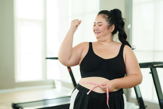 Chubby Asian woman exercise in gym, Raise hands in joy progress with tape measure, triumph in health and fitness journey. Elated female measuring waist, a victorious moment in her workout regimen