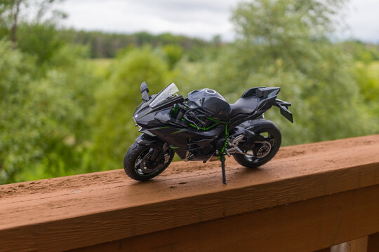 A miniature model of a black sports motorcycle - perched on a wooden railing - overlooks a lush green landscape - cloudy sky. Taken in Toronto, Canada.