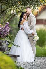 Wedding portrait of smiling newlyweds. A stylish groom in a beige suit and a cute brunette bride in a white dress are tenderly embracing in the park