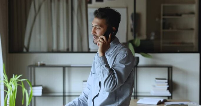 Positive young Indian project man leader talking on mobile phone in office, speaking on cell, smiling, discussing startup, business communication on telephone call, looking away, smiling