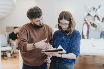Two young business professionals discussing notes in a busy office workspace.