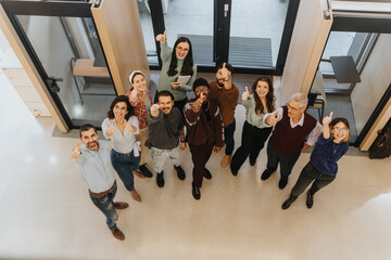 Overhead view of a multicultural team smiling and showing thumbs up in an office setting,...