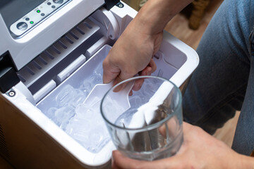 Closeup shot of a person's hand transferring bullet type ice cubes from a small portable ice maker into a clear glass with a small scoop.