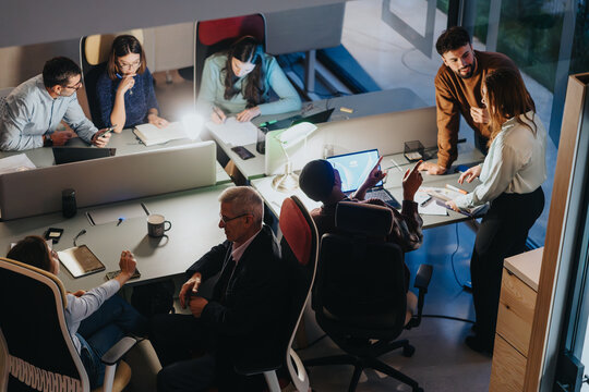 Overhead view of a diverse business team working late, collaborating around a table in a modern office setting.