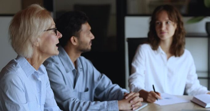 Senior blonde business professional woman talking to listening nodding younger colleagues at corporate meeting, sharing ideas for teamwork, brainstorming. Diverse team discussing cooperation