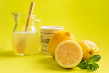 Several lemons with one cut in half and mint, in the blurred background a jug with lemonade and a jar of sugar, greenish background.