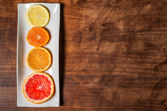 Rectangular white plate with slices of various citrus fruits, grapefruit, orange, lemon and tangerine, on a dark wooden table, with copy space, overhead view.