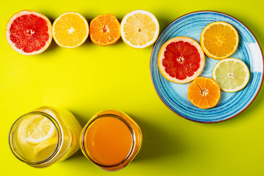 Slices of grapefruit, orange, lemon and tangerines on a plate along with half fruits and two jars with juices, greenish background, top view.
