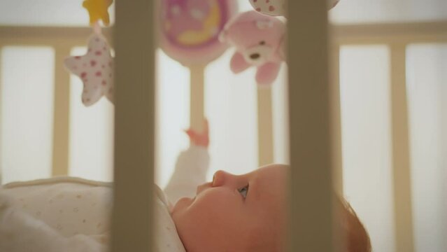 Girl. A newborn child in a bodysuit carefully reaches out to toys in a crib. Cinematic shot of a young mother paying attention to her beloved child, modern parenting, parent