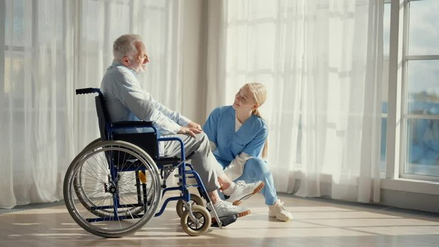Nurse talking senior man in wheelchair. Female social welfare physical therapist looks after senior patient, listening, provide medical care at home. Living with legs paralysis, special needs people.