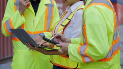 Three workers wearing hard hats are using tablets working together in a container yard, a shipping...