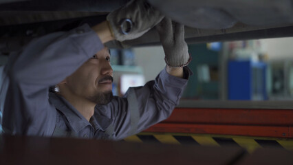 A man is working on a car under a lift. He is wearing a grey uniform and a pair of gloves