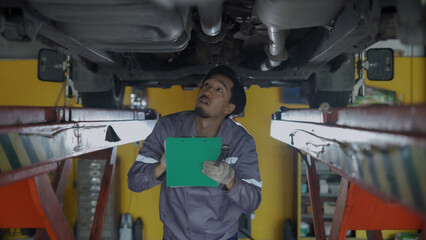 A man is working on a car under a lift. He is writing on a clipboard. Scene is serious and focused