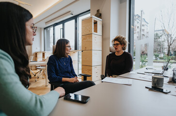 Three businesswomen conducting a business meeting in a bright office space with laptops, papers,...