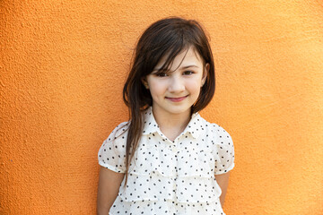 Front portrait of a cheerful shy little girl in dress posing near orange wall, looking at camera....