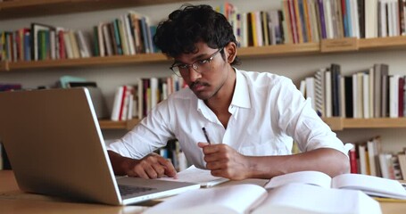 Focused Indian student guy sit at desk in library writing essay, making notes in workbook, learning theory, gaining new knowledge, e-learning using computer application. Modern tech, education, skills