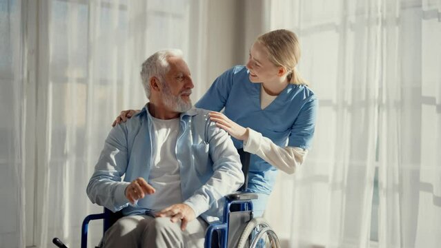 Social worker helps man in wheelchair. Woman nurse social welfare physical therapist looks after senior male, talking, providing medical care at hospital. Living with paralysis, special needs people.