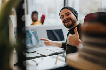 Cheerful bearded man giving thumbs up in a casual office setting, signifying approval or success with laptops in background. Positive results concept.