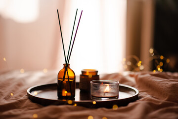 Liquid home perfume in dark brown glass bottle and bamboo sticks with burning candles on wooden tray in bed room close up. Cozy hygge atmosphere.