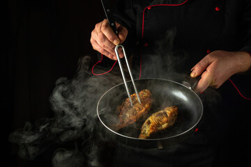 The cook fries carp fish in a frying pan in a restaurant kitchen. Idea of a delicious fish diet on a black background. Fork in the chef hand