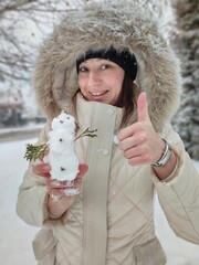 Smiling woman with the fur hood holding little snowman figure and giving thumb up.