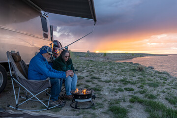 Matured Caucasian couple warming their hands by a campfire sitting under an awning by a lake at...