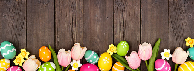 Easter eggs and flower decorations. Top view bottom border against a dark rustic wood banner background. Copy space.