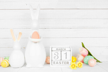 Easter decor on a white shelf against a white wood panel background. Cute ceramic bunnies,...