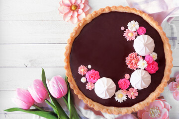 Delicious chocolate tart with meringue and pink chocolate flowers. Top down view table scene on a white wood background. Spring baking concept.