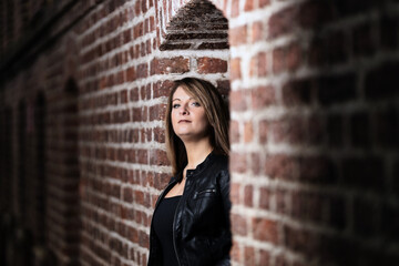 Woman model around thirty in black leather jacket and hot pants in front of urban brick wall.
