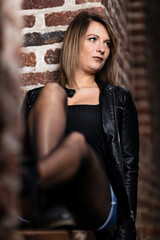 Woman model around thirty in black leather jacket and hot pants in front of urban brick wall.