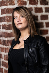 Woman model around thirty in black leather jacket and hot pants in front of urban brick wall.