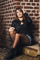 Woman model around thirty in black leather jacket and hot pants in front of urban brick wall.