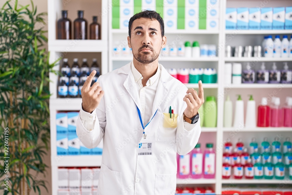 Poster Handsome hispanic man working at pharmacy drugstore pointing up looking sad and upset, indicating direction with fingers, unhappy and depressed.