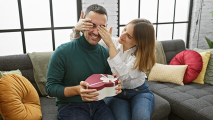 A woman surprises a man with a heart-shaped gift box, hinting at romance in a cozy living room...